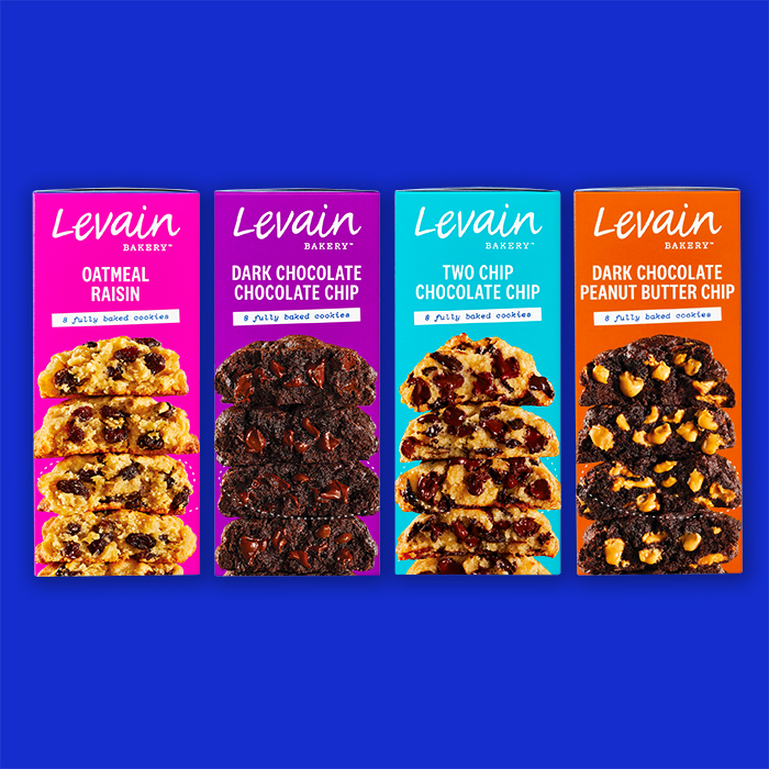 Four side panel facings of Levain Bakery Frozen Cookie boxes on a blue background. Each colorful side panel features the Levain Bakery logo, cookie flavor, "8 fully baked cookies" descriptor, and a photograph of the flavor's 4 cookies stacked. In order from left to right, a bright pink side panel for Oatmeal Raisin, a purple for Dark Chocolate Chocolate Chip, a light cyan for Two Chip Chocolate Chip, and a deep orange for Dark Chocolate Peanut Butter Chip.