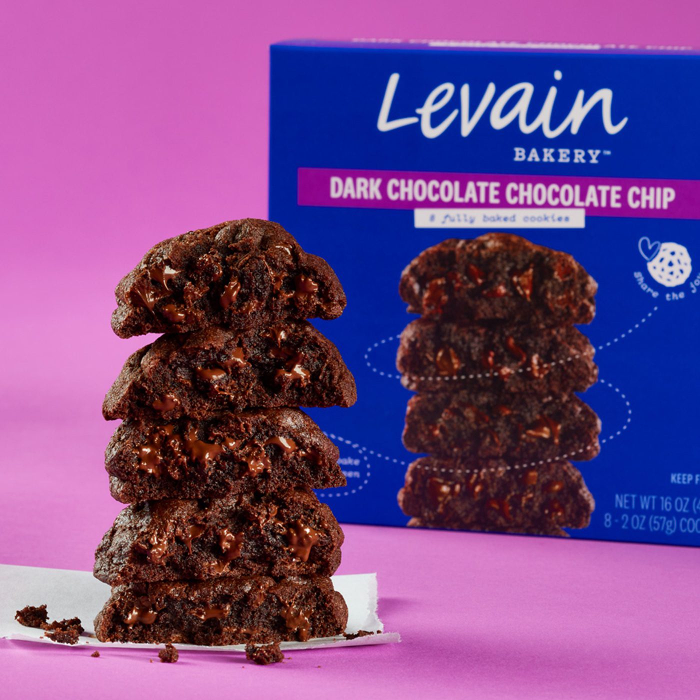 An open stack of 5 Levain Bakery Dark Chocolate Chocolate Chip cookies, revealing a gooey chocolate center, photographed on a purple-colored background. Featured next to the cookie stack is a blue retail box of Levain Bakery Dark Chocolate Chocolate Chip Frozen Cookies.