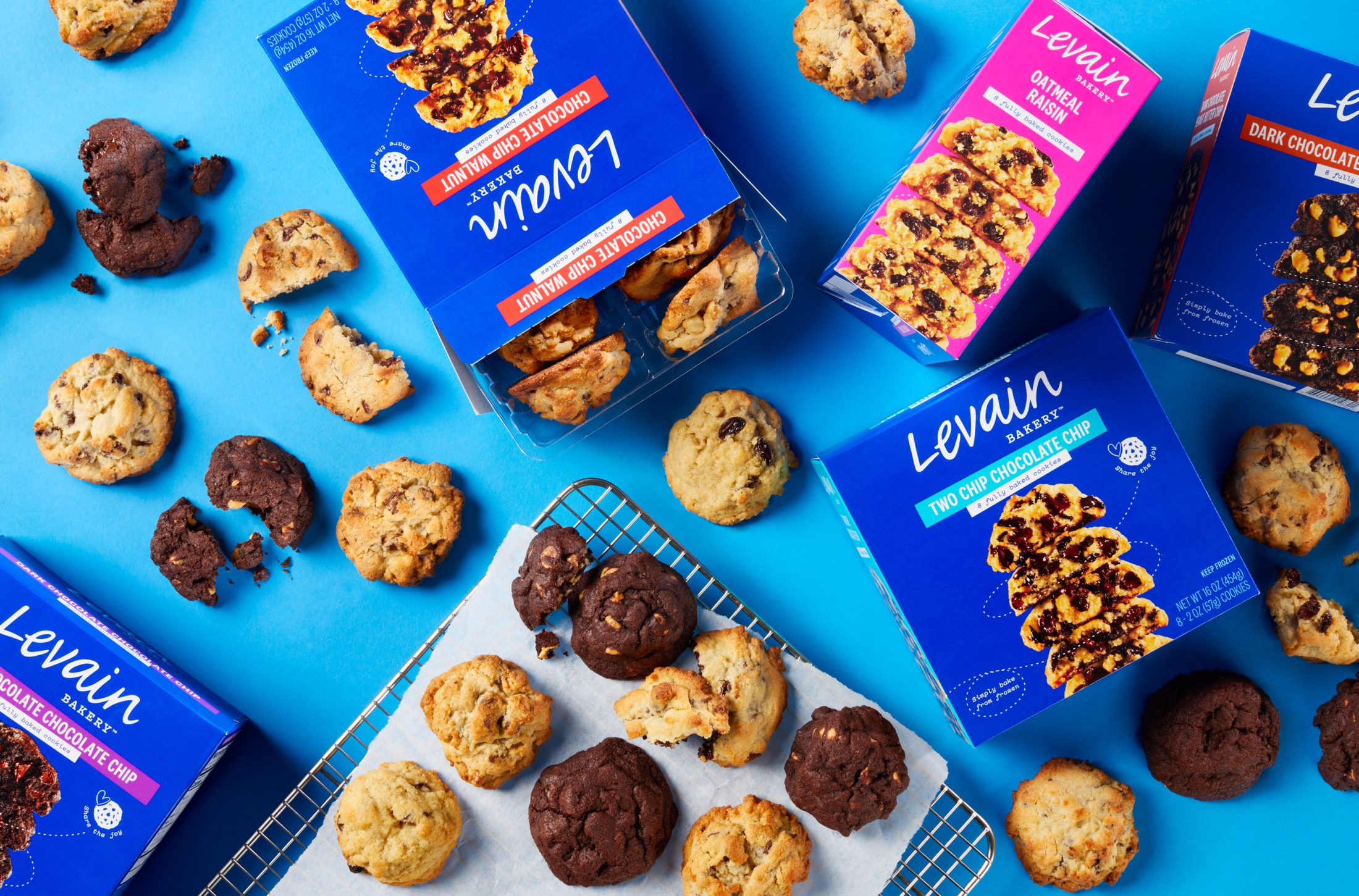 New range of Levain Bakery Fully Cooked Frozen Cookie boxes displayed on a blue background. One box opened displaying plastic tray of cookies with additional cookies on a baking rack, some cookies broken in half to view gooey center. Flavors include Chocolate Chip Walnut, Oatmeal Raisin, Two Chip Chocolate Chip, Dark Chocolate Chocolate Chip, and Dark Chocolate Peanut Butter Chip.