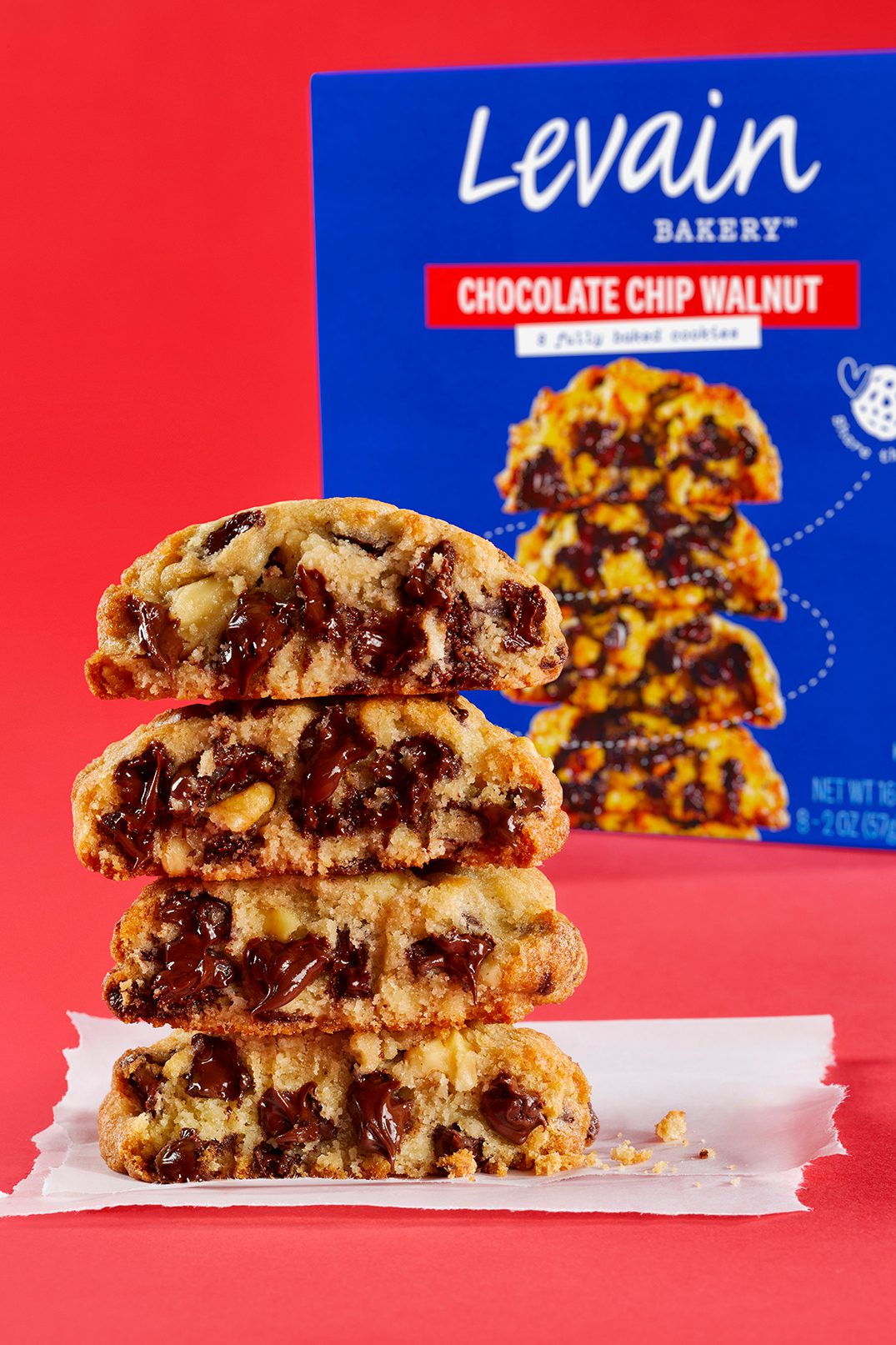 An open stack of 4 Levain Bakery Chocolate Chip Walnut cookies, revealing a gooey chocolate center, photographed on a red-colored background. Featured next to the cookie stack is a blue retail box of Levain Bakery Chocolate Chip Walnut Frozen Cookies.