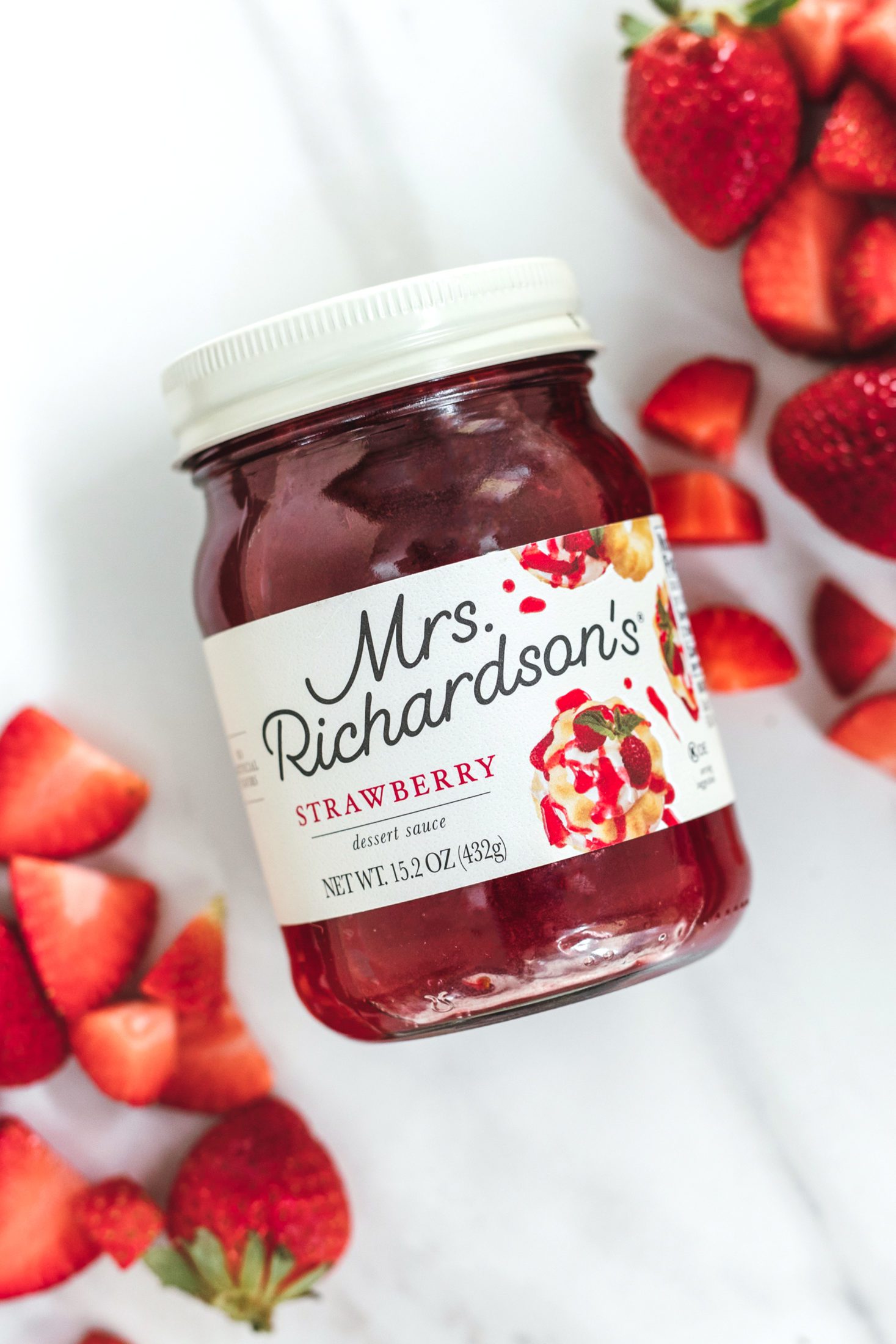 An overhead view of new Mrs. Richardson's cream label design using a clear glass jar with cream white lid. Strawberry dessert sauce jar with chopped strawberries on a marble surface.