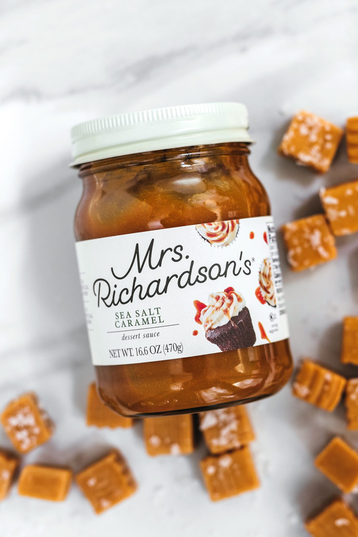 An overhead view of new Mrs. Richardson's cream label design using a clear glass jar with cream white lid. Sea Salt Caramel dessert sauce jar with sea salt caramel squares scattered on a marble surface.