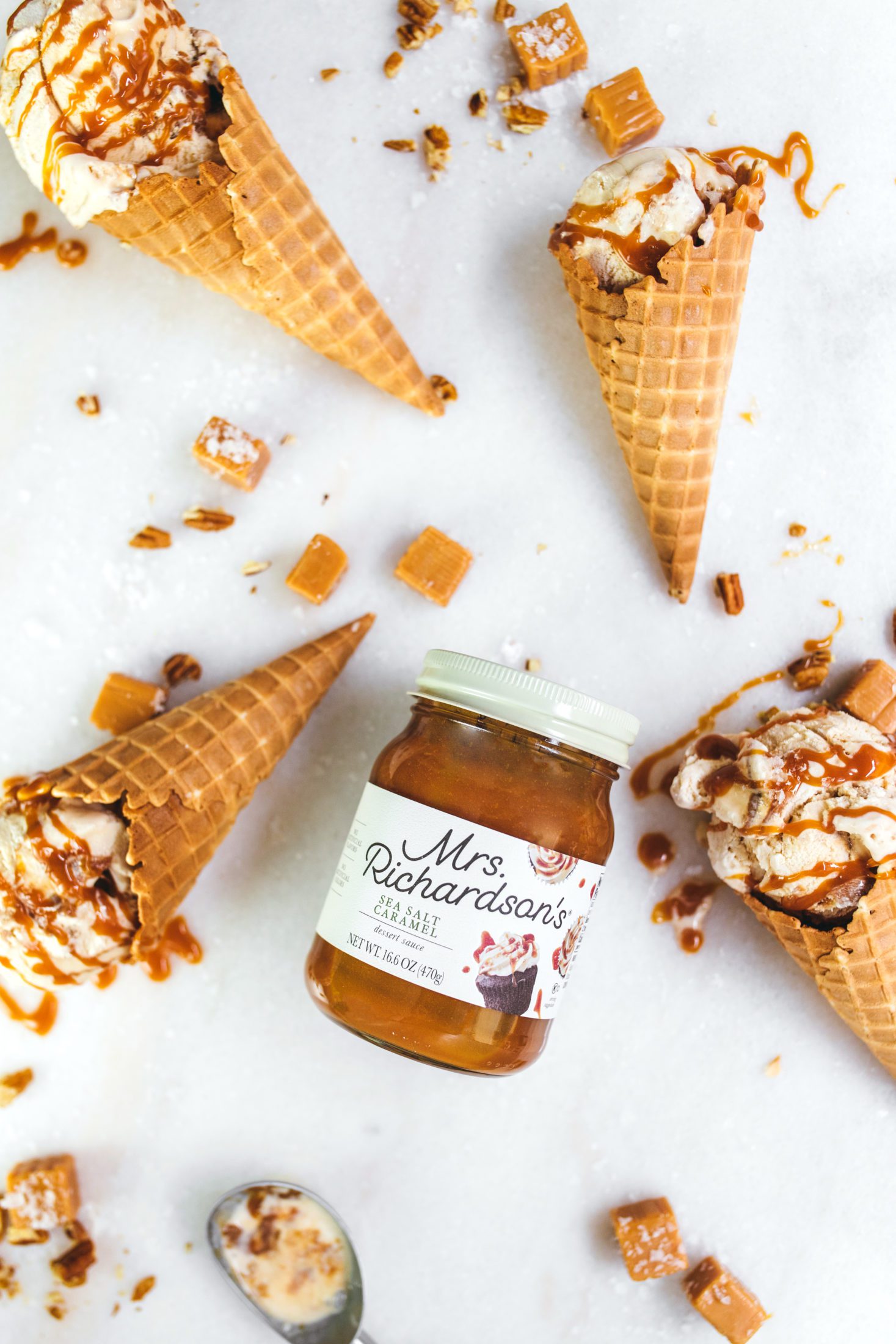 An overhead view of Mrs. Richardson's Sea Salt Caramel dessert sauce jar surrounded by sea salt caramel ice cream waffle cones and pieces of caramels on a marble surface.