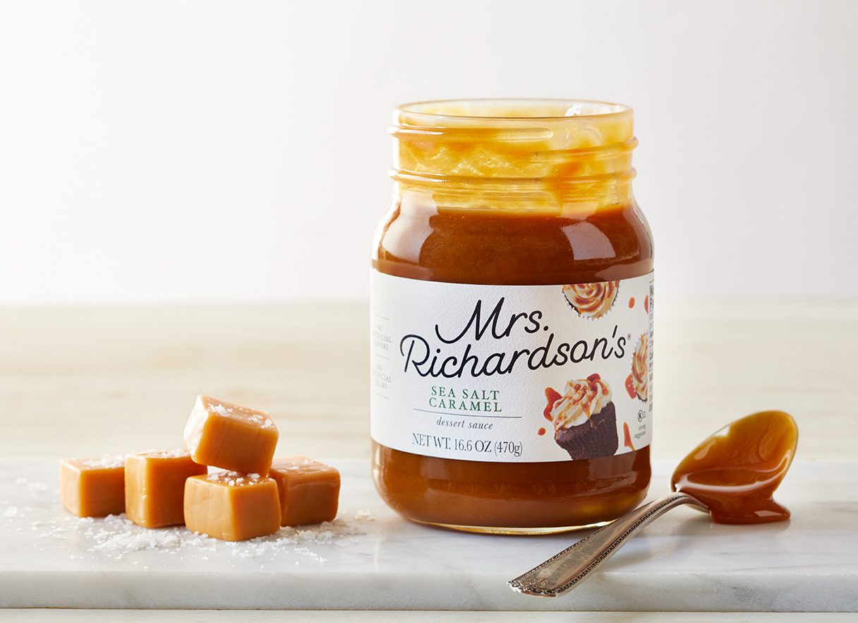 An opened jar of Mrs. Richardsons's Sea Salt Caramel dessert sauce with a dripping spoonful of caramel and caramel squares sprinkled with sea salt, all on a marble surface tabletop.