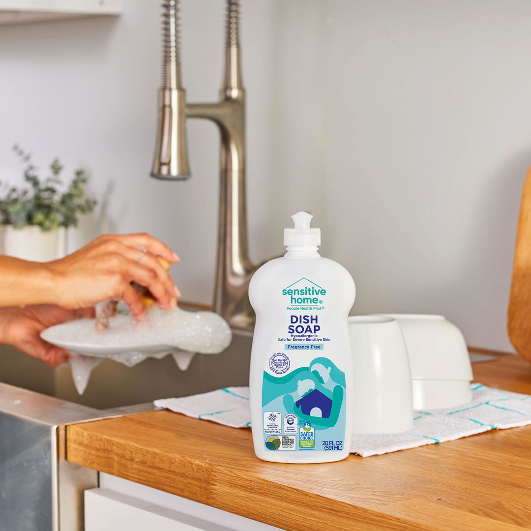 A bottle of Sensitive Home Dish Soap on a wooden kitchen counter next to the sink and some dishes. In the background, a pair of hands are washing a soapy plate over the sink.