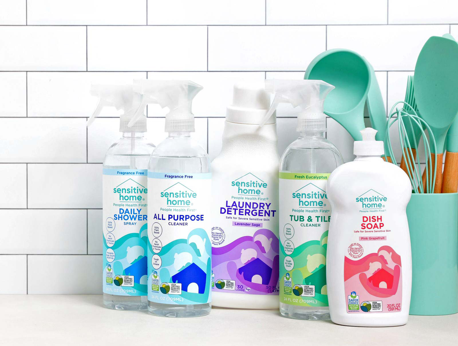 A small lineup of Sensitive Home products featuring spray bottle cleaners, dish soap, and laundry detergent photographed on a kitchen counter with white subway tile walls and kitchen utensils in the background.