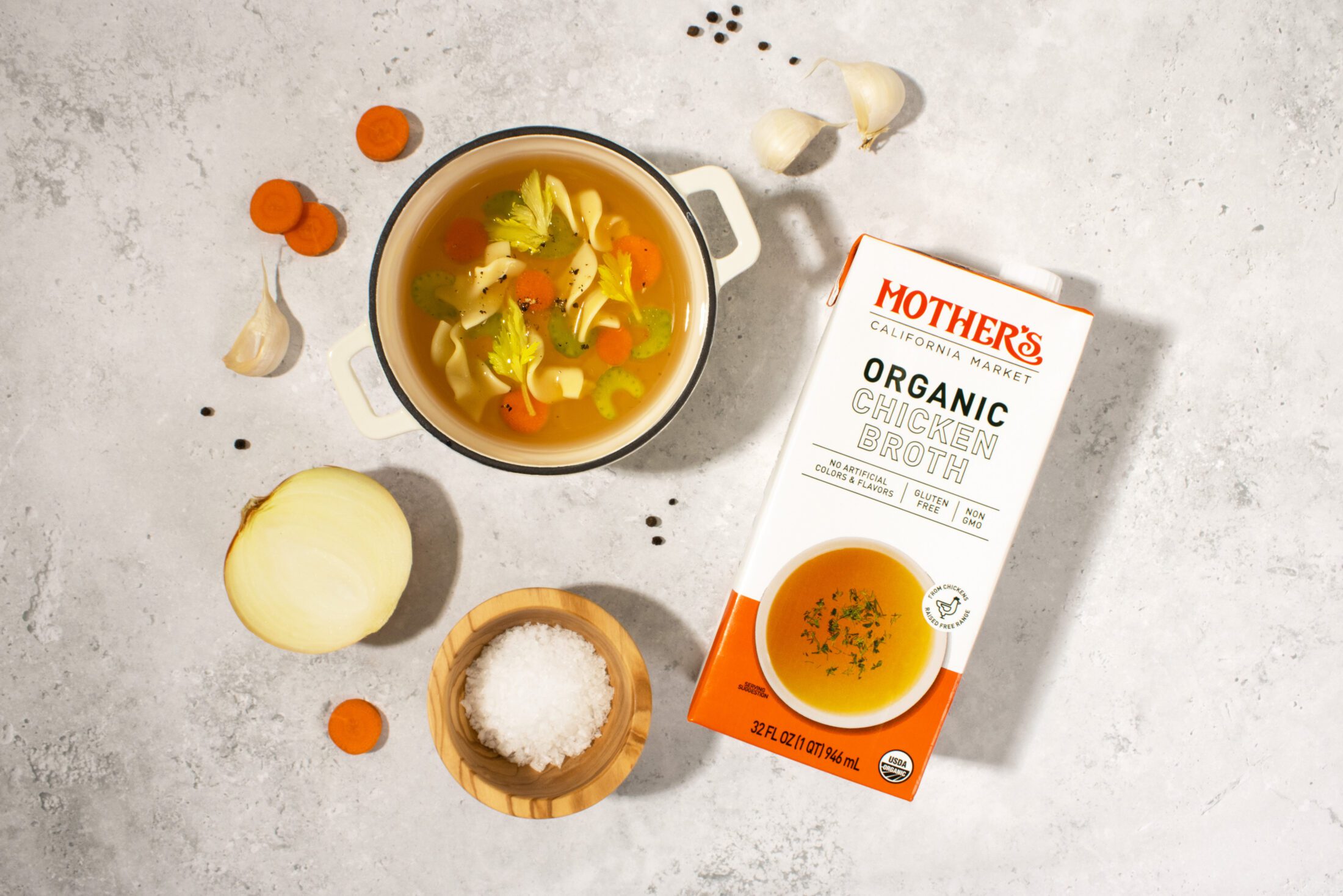 Mother's Market Organic Chicken Broth tetrapak carton laid on a white gray stone surface with a bowl of chicken noodle soup and surrounding scattered ingredients of carrot slices, half an onion, garlic cloves, and black peppercorns.
