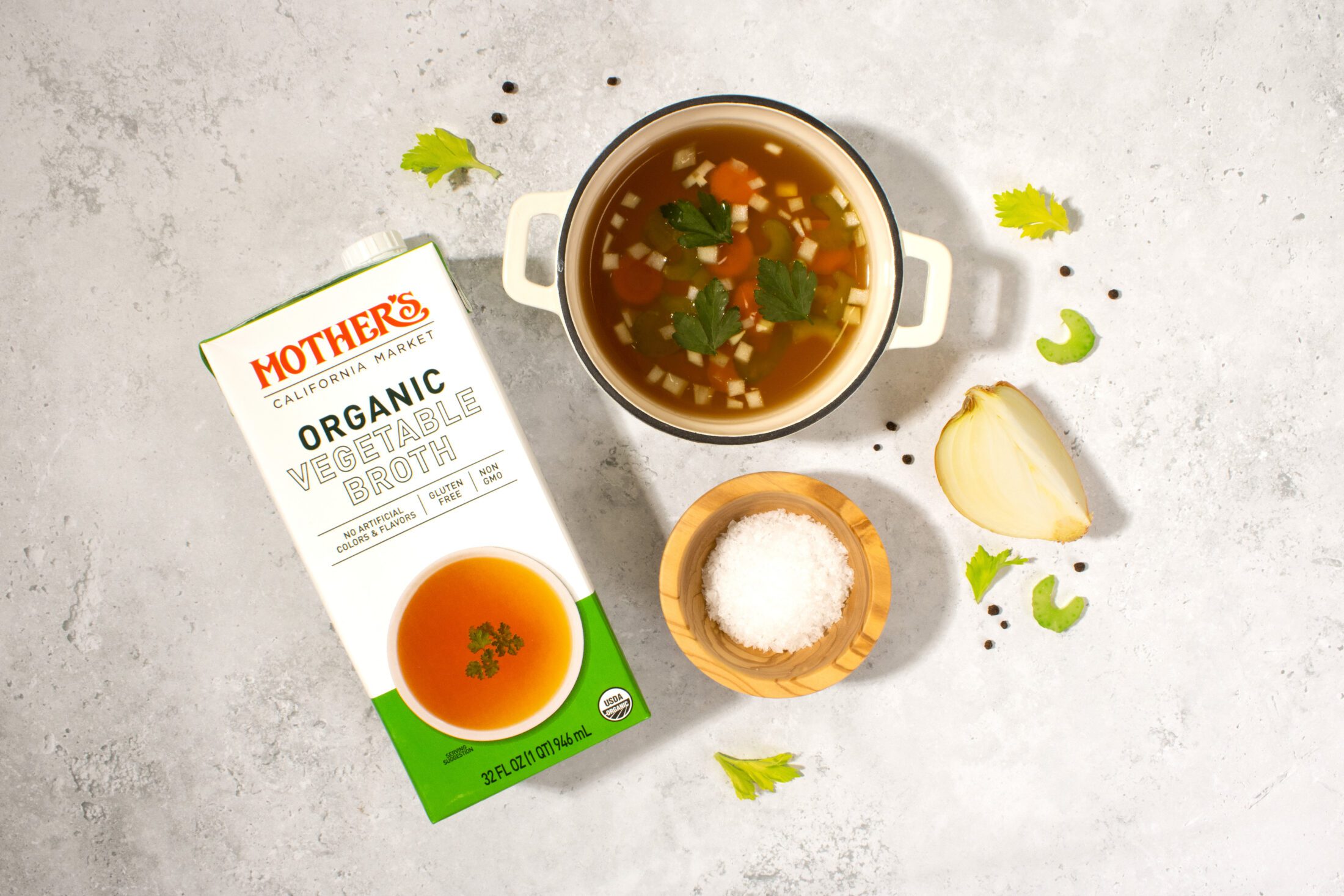 Mother's Market Organic Vegetable Broth tetrapak carton laid on a white gray stone surface with a bowl of vegetable soup and surrounding scattered ingredients of sliced celery pieces, quarter of an onion, a wooden bowl of salt, and black peppercorns.