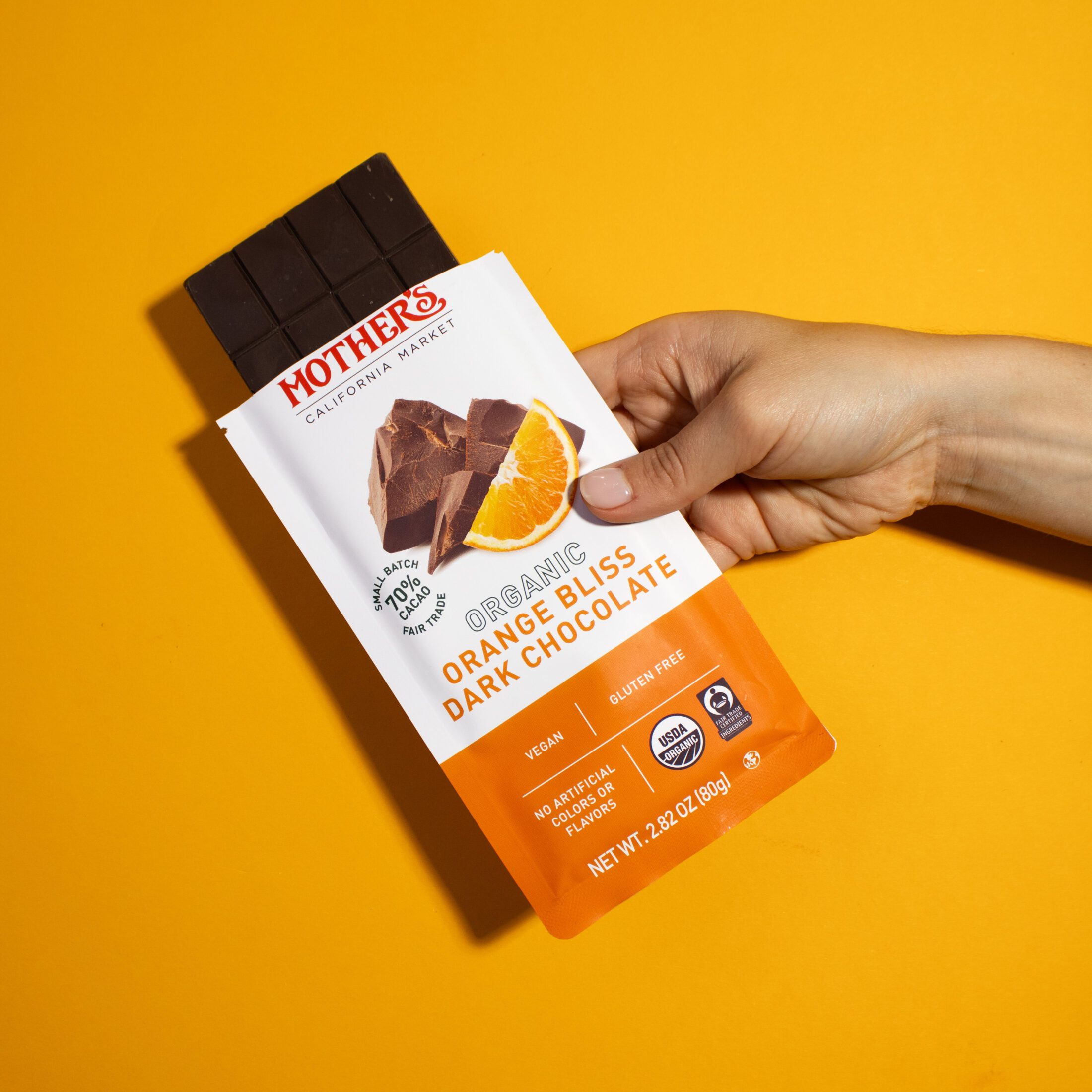 A hand holding an opened bar of Mother's Market Organic Orange Bliss Dark Chocolate in front of a bright orange backdrop.