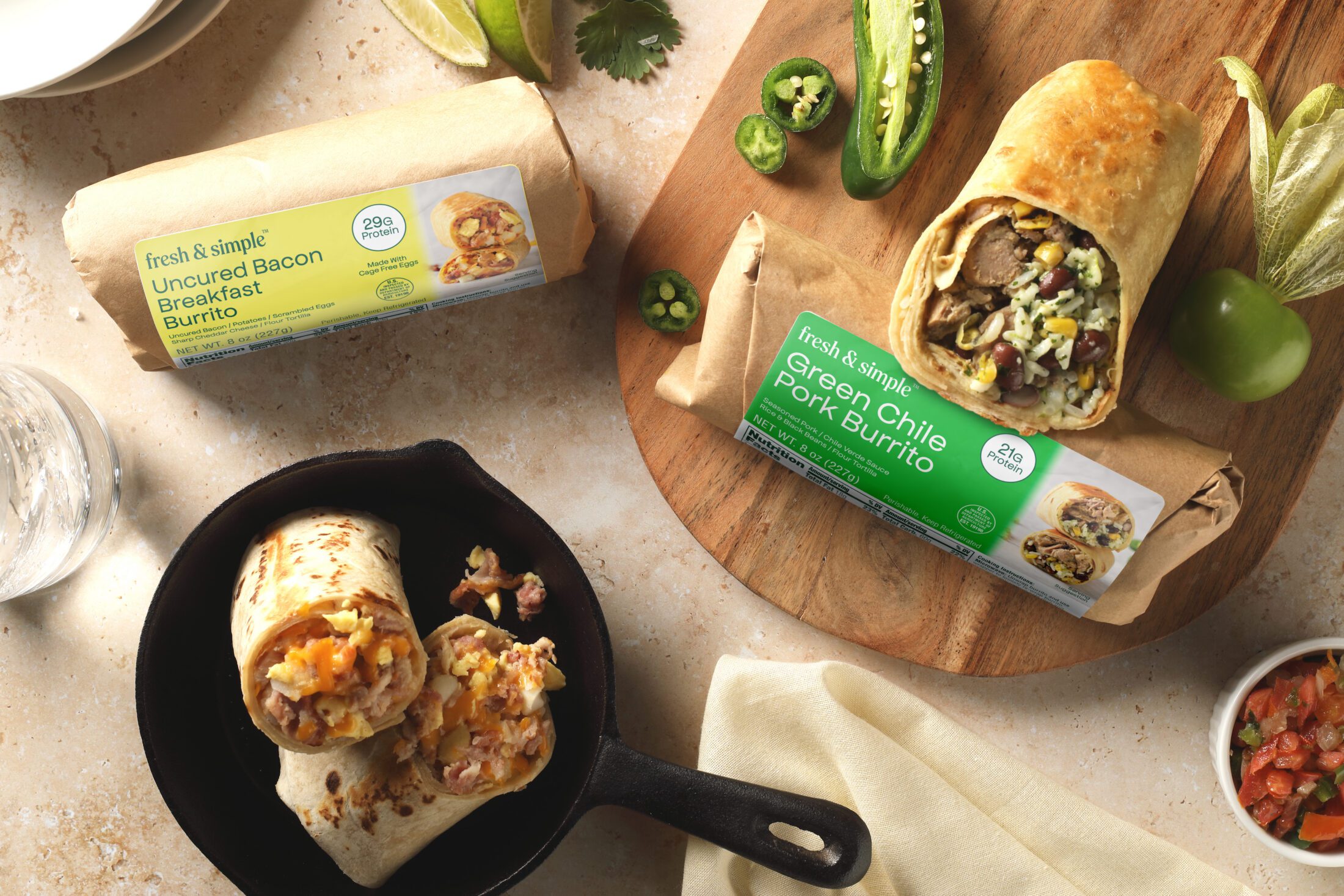 Two Fresh & Simple burritos overhead. On the left is the Uncured Bacon Breakfast Burrito in yellow packaging. Below is that burrito in a cast iron skillet cut in half. Two the right on a cutting board is the Green Chile Pork Burrito in its green packaging. Half of that burrito sits on top of the packaging.
