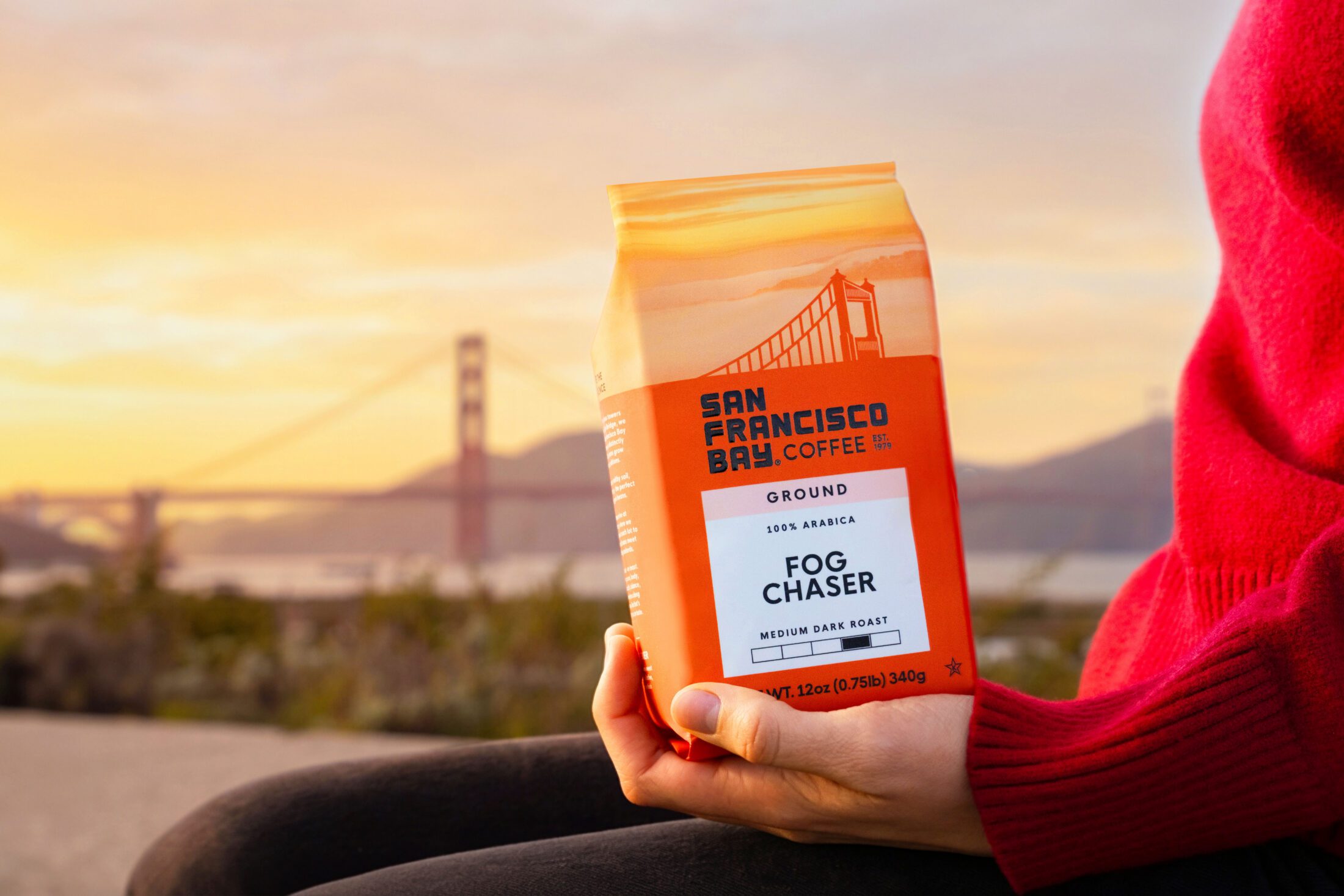 A women holding a bag of San Francisco Bay Coffee Fog Chaser Ground 28 oz infront of the the Golden Gate Bridge During Golden Hour
