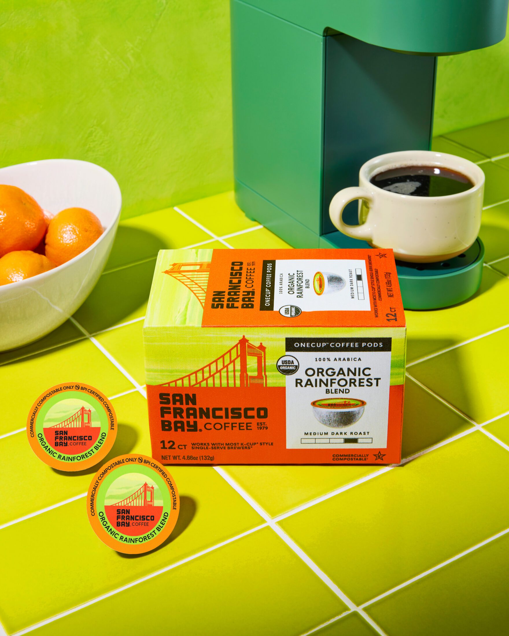 A box of San Francisco Bay Coffee Organic Rainforest Blend 12 count OneCups sits next two two individual pods in front of a freshly made coffee in the green OneCup coffee machine