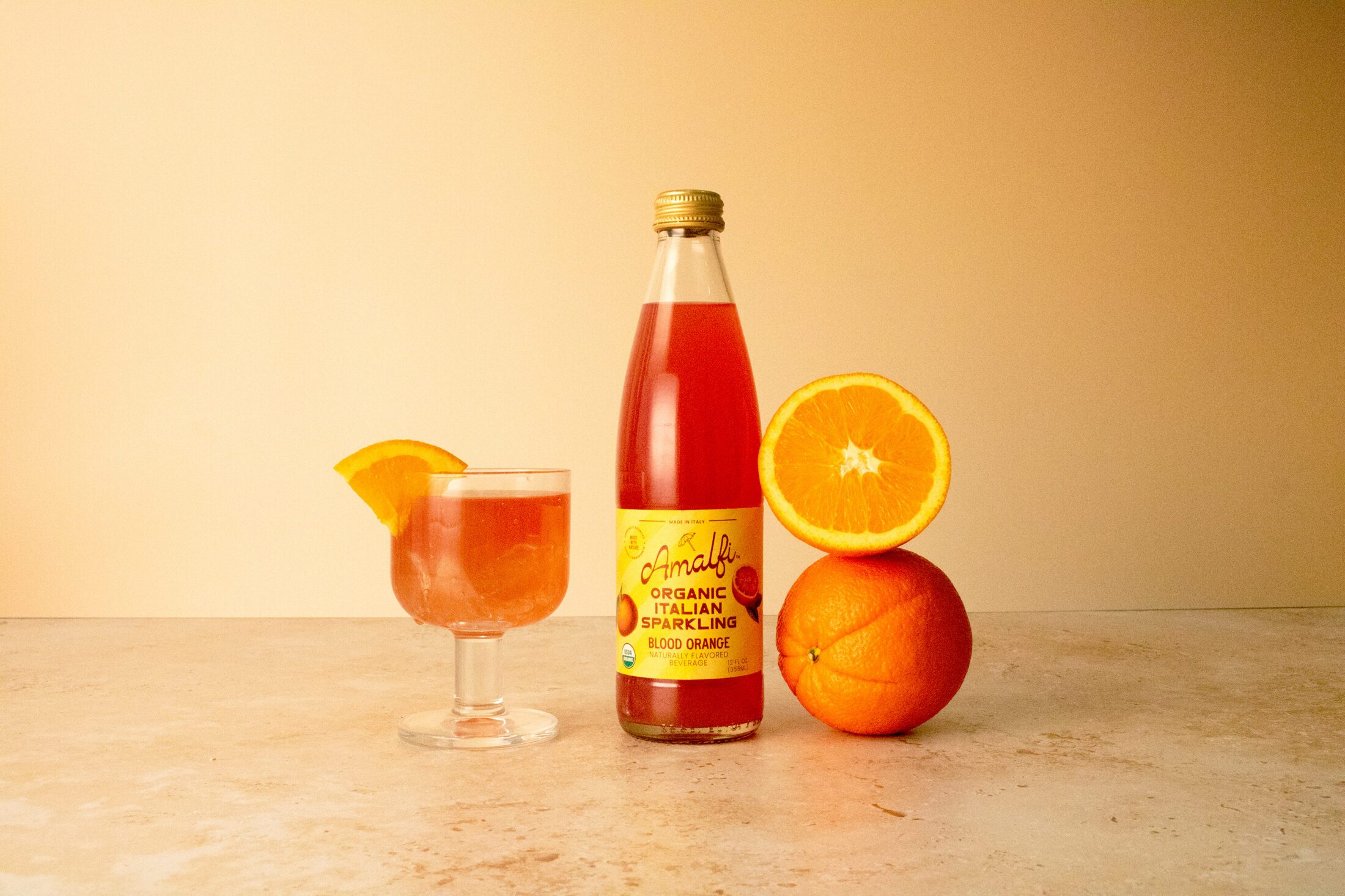 A bottle of Organic Amalfi Blood Orange Italian Soda next to a glass featuring the Italian soda as well as stacked oranges.