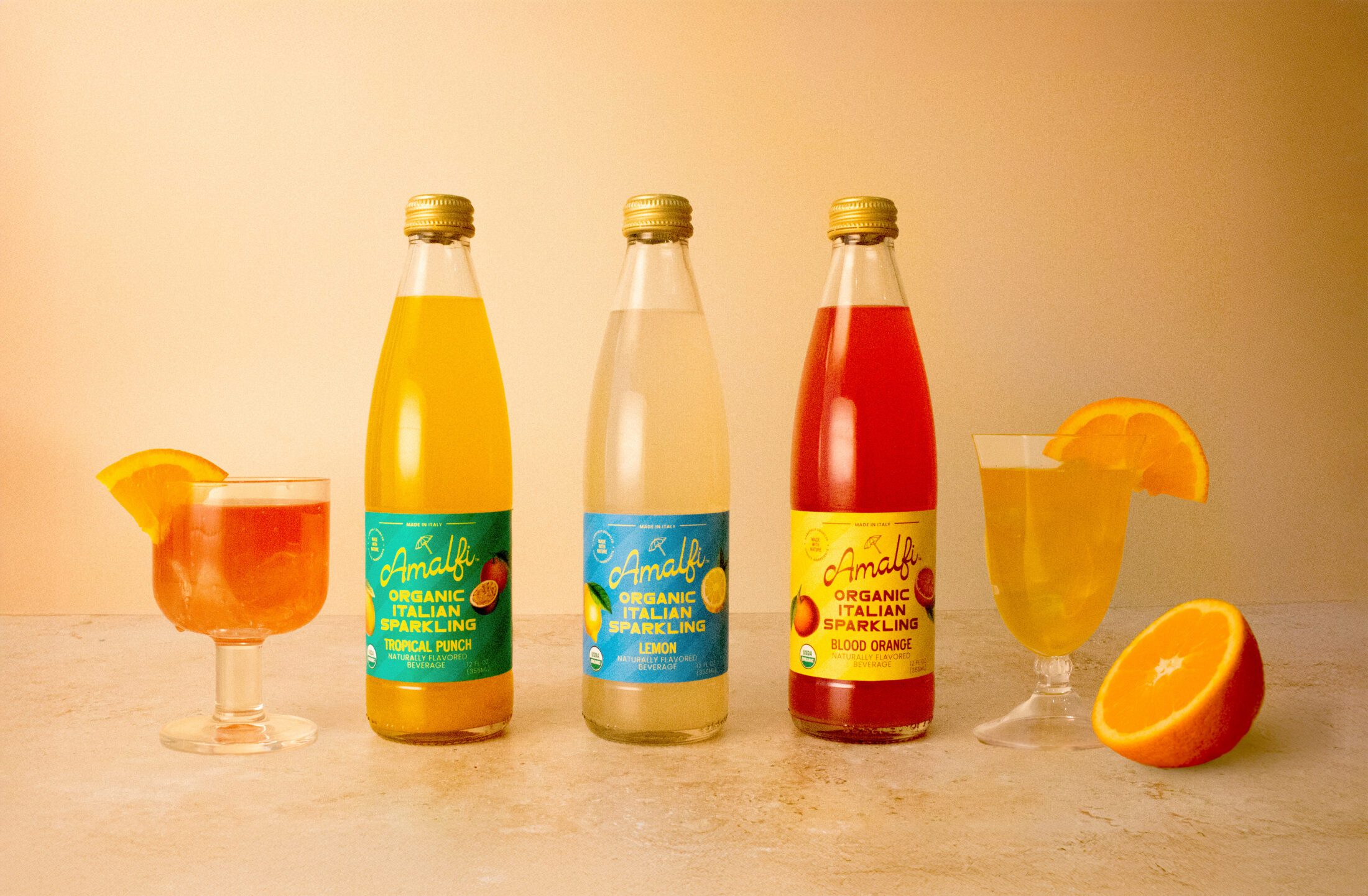Three bottles of Organic Amalfi Italian Soda - Tropical Punch, Lemon and Blood Orange with two glasses filled glasses and half an orange.