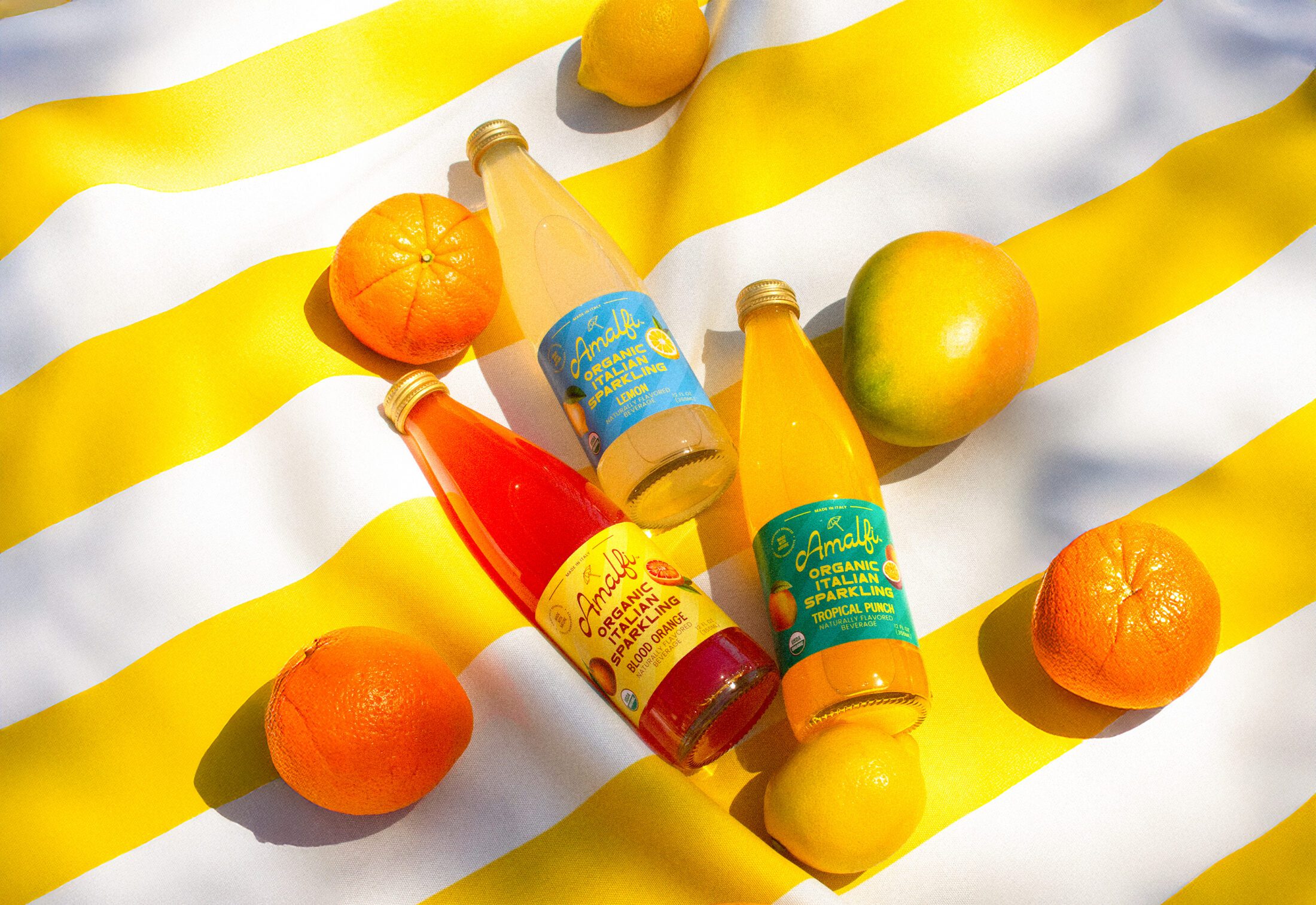 Three bottles of Amalfi Italian Soda - Tropical Punch, Lemon and Blood Orange on a yellow striped beach towel surrounded by fruit.