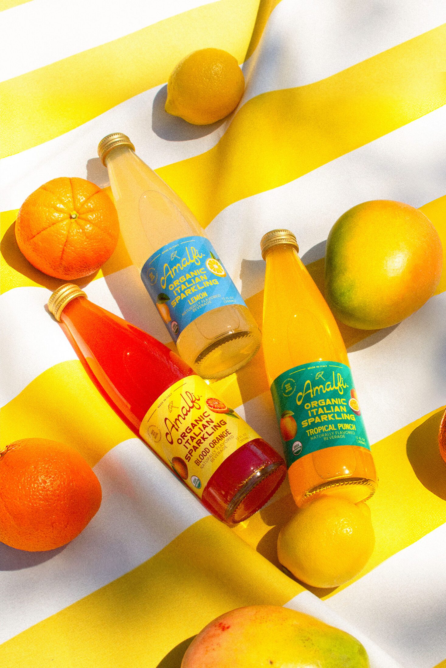 Three bottles of Amalfi Italian Soda - Tropical Punch, Lemon and Blood Orange on a yellow striped beach towel surrounded by fruit.
