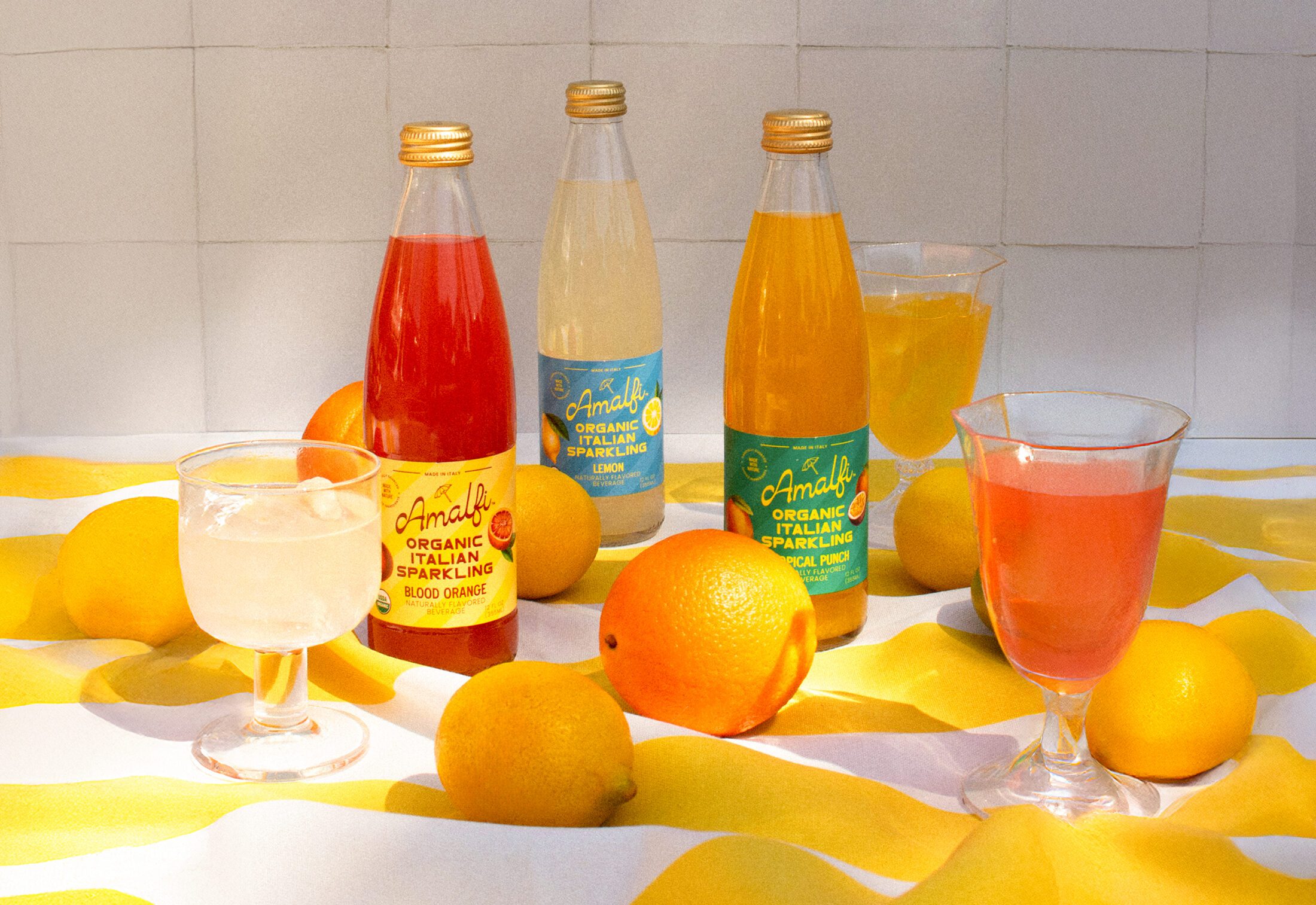 Three bottles of Amalfi Italian Soda - Tropical Punch, Lemon and Blood Orange surrounded by glasses of the soda and fruit.