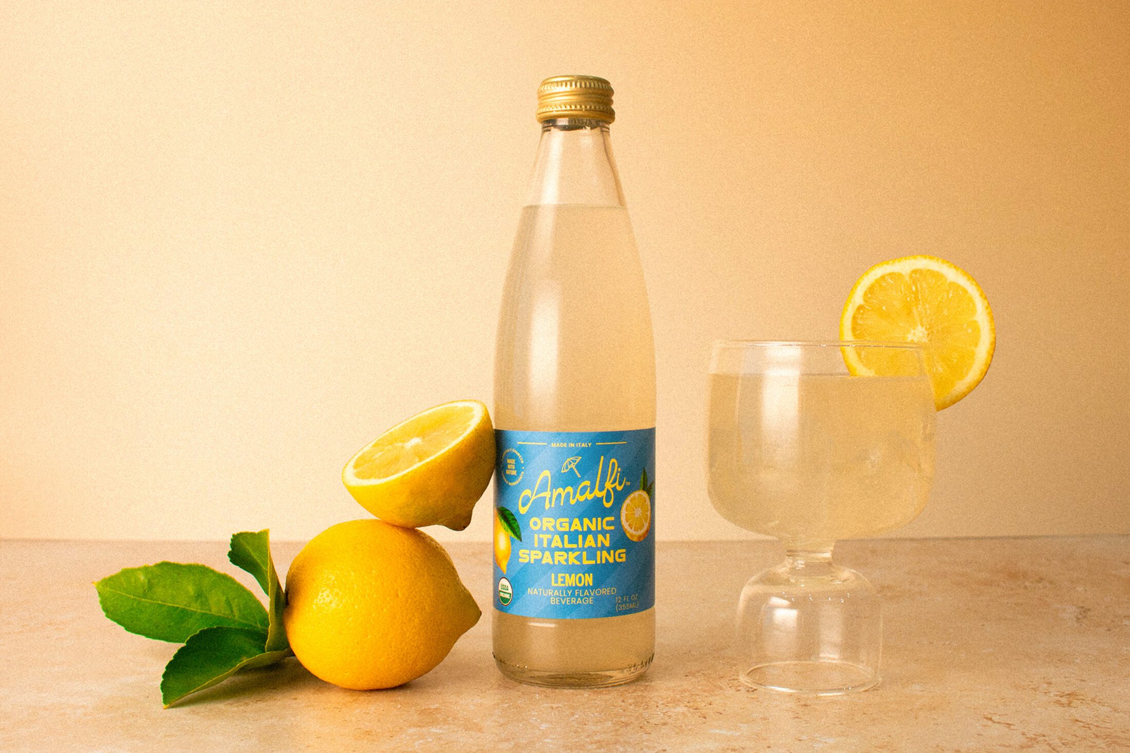 A bottle of Organic Amalfi Lemon Italian Soda next to a glass featuring the Italian soda as well as stacked lemons.