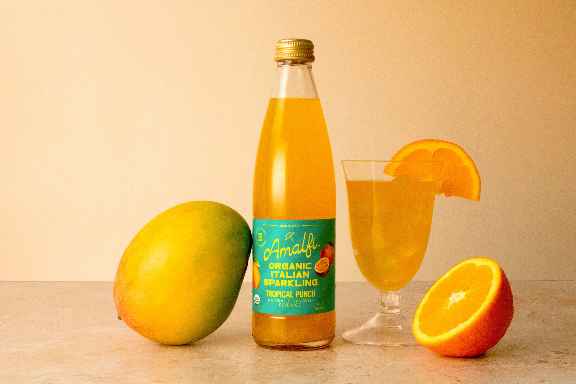 A bottle of Organic Amalfi Blood Orange Italian Soda next to a glass featuring the Italian soda as well as a mango and half an orange.