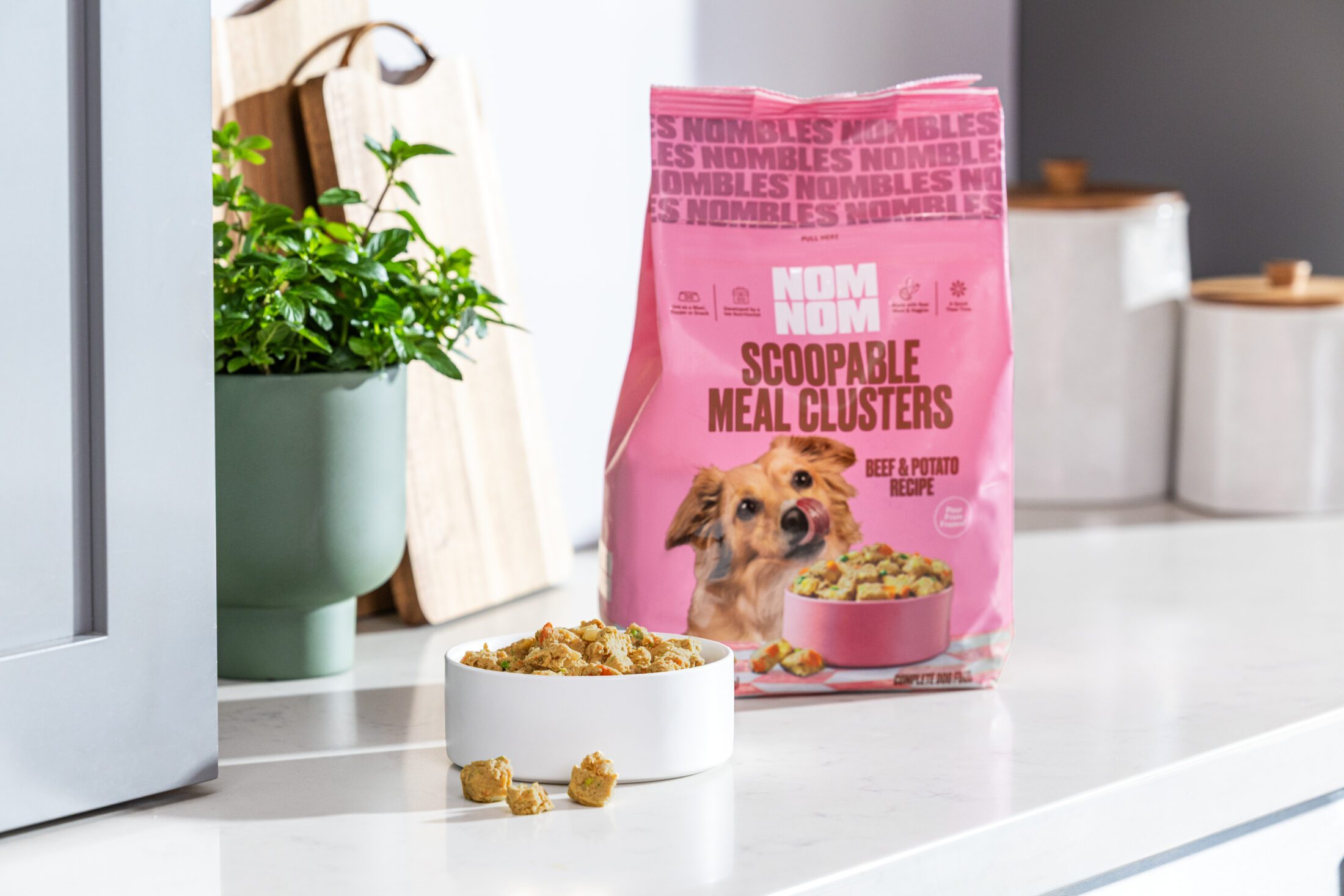 A white dog bowl with a heaping pill of Nom Nom Scoopable Meal Clusters in it. A couple pieces sit outside the bowl and in front of it. Behind the bowl is a bag of the Beef & Potato Recipe in Chew Toy Pink.