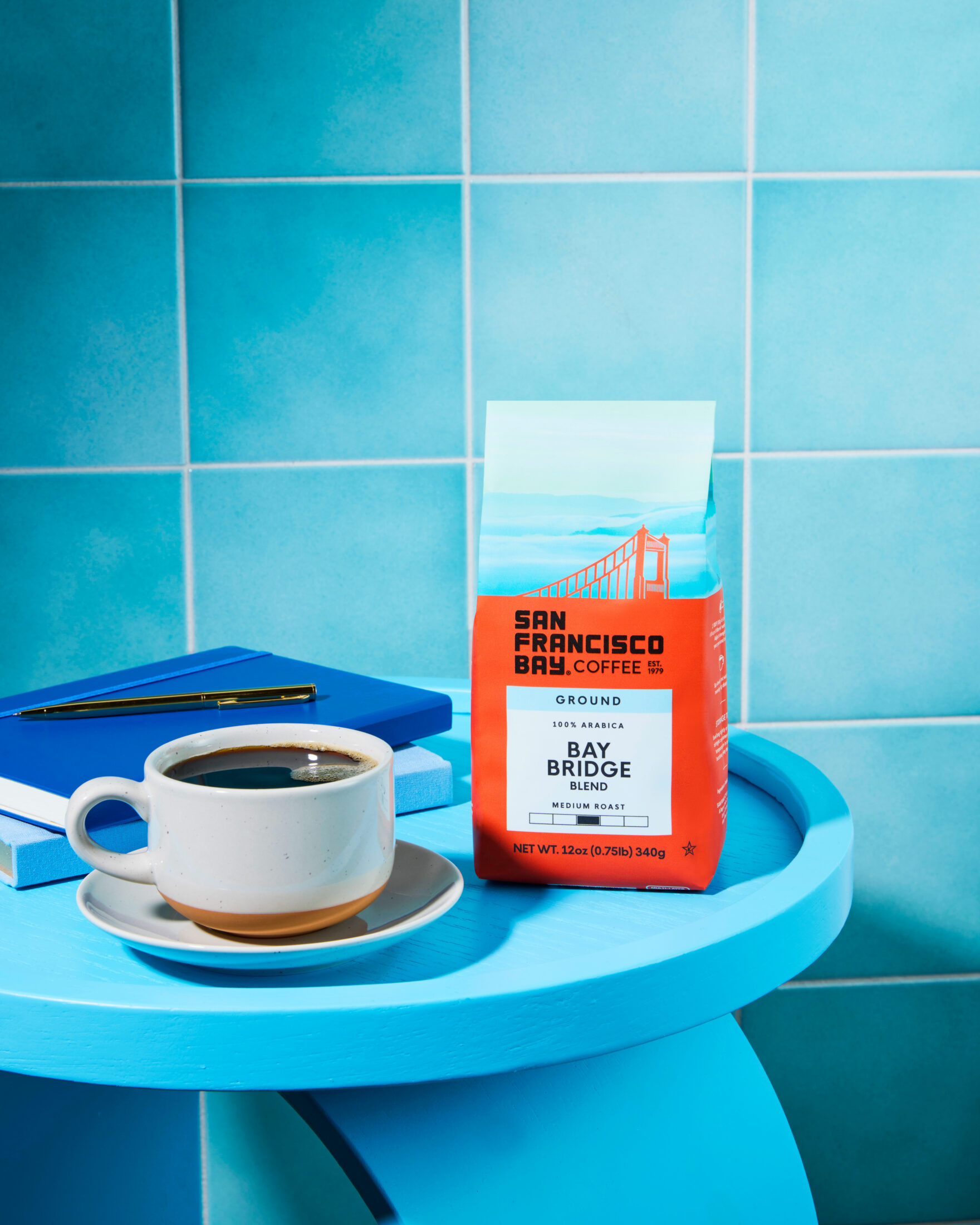 San Francisco Bay Coffee Bay Bridge Coffee 12oz Bag sitting on top of a blue side table next to a cup of coffee