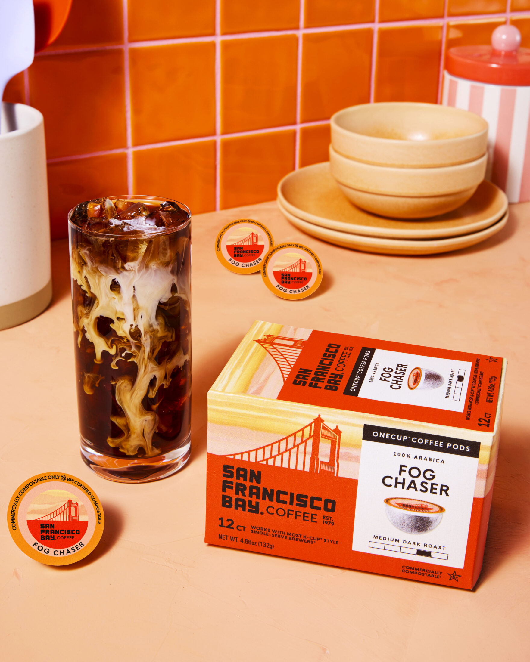 San Francisco Bay Coffee Fog Chaser 12 Count OneCup box next to a cup of iced cofee with milk on top of an orange surface that matches the coloring of the box